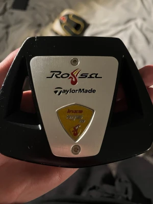 Taylor Made Putter - En svart och silverfärgad golfputter från TaylorMade, modell Rossa. Klubban har en robust design med metallplatta framtill och detaljer i rött och gult. Greppet är svart med röda och gula inslag. Perfekt för golfentusiasten.