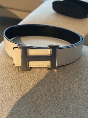 Hermess vitt skärp med H-spänne - Snyggt vitt skärp från Hermess med ett ikoniskt H-formad spänne i silver. Skärpet är tillverkat i skinn och har en stilren, bred design som ger ett lyxigt intryck. Perfekt för att lyfta din outfit med en exklusiv touch.