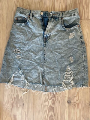 Ljusblå jeanskjol från Divided - Trendig ljusblå jeanskjol från Hm med slitna detaljer och fransig nederkant. Kjolen har klassisk femficksdesign, knapp och dragkedja 