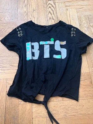 Svart BTS t-shirt med knyt - Svart t-shirt med stort holografiskt  , dekorativa metalldetaljer på axlarna och knytning nedtill. Perfekt för dig som gillar K-pop och vill ha en cool och avslappnad stil.