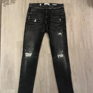 Replay Anbass svarta skinny jeans - Svarta Replay Anbass jeans med slitna detaljer och skinny passform. Jeansen har klassisk femficksdesign, dragkedjegylf och tydliga slitningar framtill för en cool, använd look. Märkeslapp på fickan och bak. Perfekta för dig som gillar streetstyle.
