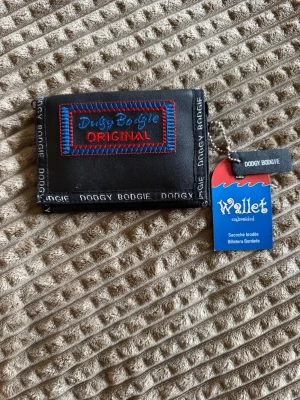 Svart plånbok Doggy Bodgie - Cool svart plånbok från Doggy Bodgie med broderad patch framtill i blått och rött. Plånboken är tillverkad i PVC med nylonfoder och har dragkedjefack på baksidan. Snygga detaljer med text längs kanterna och praktisk storlek för fickan.