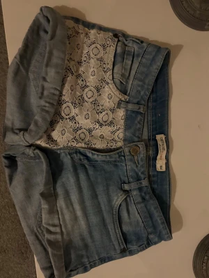 Blå jeansshorts med spetsdetaljer - Snygga blå jeansshorts från Lindex med vit spets på ena framsidan och bakfickan. . Perfekta för en trendig sommarlook! Strl 164, (xs) midjemått se bild