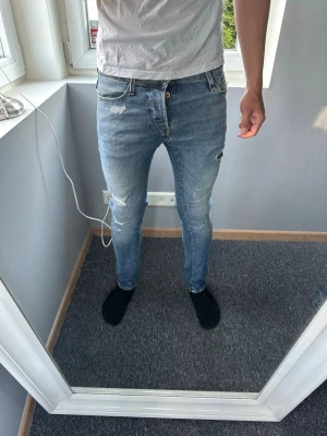Jack&jones jeans med slitningar - Säljer ett par ljusblå skinny jeans med slitningar och hål på låret. Jeansen har klassisk femficksdesign, låg midja och dragkedjegylf. Perfekta för en avslappnad och trendig look.