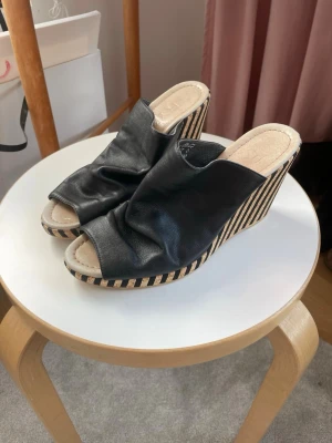 Svarta kilklackade sandaler - Snygga svarta sandaler med öppen tå och kilklack. Ovandelen är i mjukt svart läderliknande material och klacken har ett randigt mönster i beige och svart. Perfekta för att lyfta din sommarstil med en cool touch.