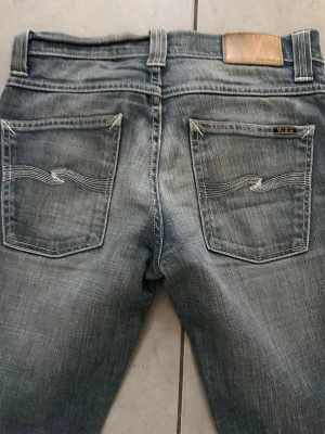 Grim tim Nudie Jeans - Snygga blå jeans från Nudie Jeans med klassisk femficksdesign och raka ben. Jeansen har ljus tvätt och dekorativa sömmar på bakfickorna. Tillverkade i bomull med en avslappnad passform som funkar till många stilar.