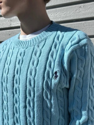 Ljusblå kabelstickad tröja Polo Ralph Lauren - Ljusblå kabelstickad tröja från Polo Ralph Lauren med rund hals och broderad logotyp på bröstet. Tröjan har ribbade muddar vid ärmslut och nederkant, och är tillverkad i mjukt material. Perfekt för dig som gillar klassisk och stilren design.