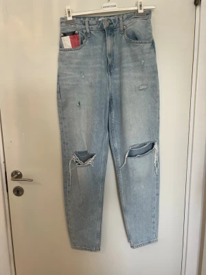 Ljusa slitna jeans från Tommy Hilfiger - Säljer ett par ljusblå mom jeans från Tommy Hilfiger med hög midja och raka ben. Jeansen har stora slitningar vid knäna och mindre slitningar framtill. Klassisk femficksmodell med Hilfiger-detaljer på ficka och bak. Storlek: W26 L32.