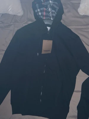 Burrberry  - Stilren svart hoodie från Burberry med hel dragkedja och klassiskt rutigt foder i huvan. Hoodien har snörning i huvan och ribbade muddar. Perfekt för dig som gillar exklusiva streetwear-plagg med snygga detaljer. Storlek S men funkar som M
