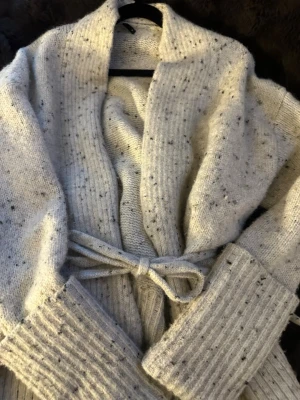 Ljusgrå stickad kofta med knytskärp - Mysig ljusgrå stickad kofta med svarta melerade detaljer och breda ribbade muddar. Koftan har ett knytskärp i midjan och en öppen, avslappnad passform. Koftan är i S men passar M.