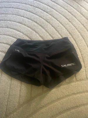 Svarta sportshorts från Salming - Säljer ett par svarta sportshorts från Salming med kort passform och elastisk midja. Materialet är lätt och snabbtorkande, perfekt för träning eller löpning. Diskret logga på ena benet och Clean Feel-tryck på andra sidan. Snygga och funktionella shorts för aktiva dagar.