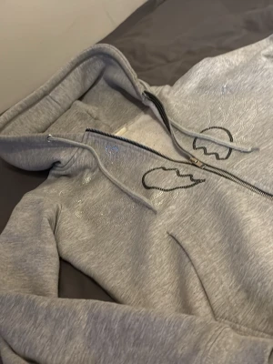 Grå hoodie med dragkedja och tryck - Snygg grå hoodie med dragkedja framtill och huva med dragsko. På bröstet finns ett tryck av ett brustet hjärta med vingar och lite glittriga detaljer. Hoodien har ribbade muddar och fickor framtill. Perfekt för en chill och avslappnad stil.