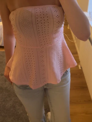Rosa axelbandslös tubtopp med broderi - Söt ljusrosa axelbandslös tubtopp med broderade hålmönster och utsvängd nederdel. Toppen har en bandeauform och är perfekt för varma dagar. Materialet känns som bomull och toppen har en feminin och trendig vibe.