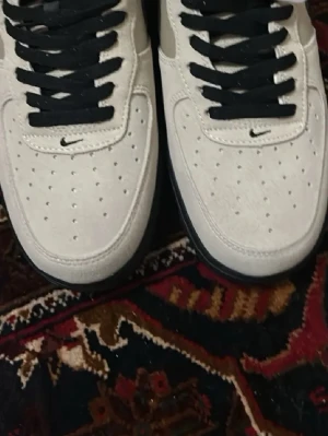 Nike Air Force 1 pris diskuterbar  - Snygga Nike Air Force 1 sneakers i beige mocka med svarta detaljer och svart sula. Klassisk låg modell med svart swoosh på sidan och svarta skosnören. Perforerad tå för extra ventilation och stilren look.