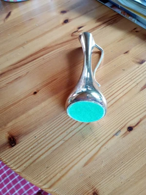 Liten silverfärgad vas med handtag - Säljer en smal och elegant vas i silverfärgad metall med ett böjt handtag. Vasen har rund botten med grön filt undertill och en smal hals som breddas upptill. Perfekt som dekoration eller för enstaka blommor.