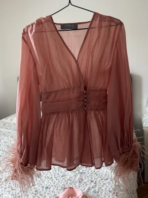 Rosa transparent blus med fjädrar - Snygg rosa blus från Varia i transparent tyg med markerad midja och knappar framtill. Långa ärmar med coola fjäderdetaljer vid ärmsluten och v-ringad hals. Finns ett linne som kommer med för att inte vara fully transparent men annars kan man ha vad man vill under. Passar xs-s