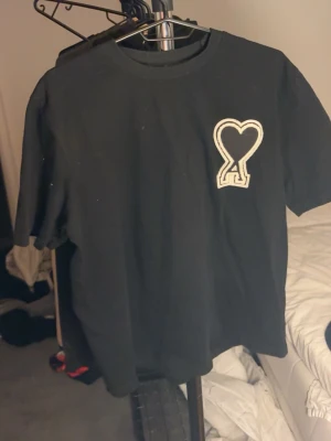 Svart t-shirt med hjärtdetalj från Ami - Svart t-shirt från Ami med korta ärmar och en stor vit broderad hjärtdetalj på bröstet. Klassisk rund halsringning och avslappnad passform. Perfekt för dig som gillar stilrena plagg med en twist.
