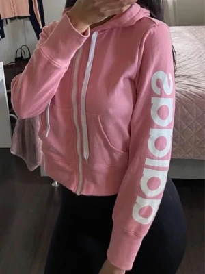 Rosa hoodie med dragkedja från Adidas - Snygg rosa hoodie från Adidas med vit dragkedja och vita snören i huvan. Stor Adidas-logga i vitt längs ena ärmen. Mjuk bomullskänsla och två fickor framtill. Perfekt för en sportig och avslappnad stil.