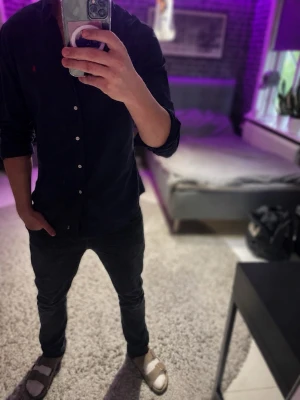 Mörkblå skjorta från Ralph Lauren - Snygg mörkblå skjorta från Ralph Lauren med klassisk krage och knappar framtill. Skjortan har en röd broderad logga på bröstet och är långärmad. Perfekt för dig som gillar stilrena och tidlösa plagg.