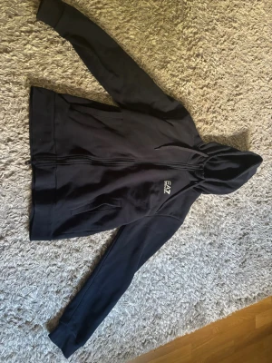 Svart hoodie från EA7 Emporio Armani - Svart hoodie med dragkedja från EA7 Emporio Armani. Klassisk design med huva och två fickor framtill. Mjuk bomullskänsla och stilren logga på bröstet. Perfekt för en avslappnad och trendig look. Det står att den är Xs men passar som M-L