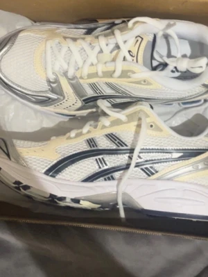 ASICS vita och beige sneakers - Snygga sneakers från ASICS i vitt och beige med svarta detaljer. Skorna har klassisk mesh-ovandel för bra andningsförmåga och silvriga inslag. Perfekta för en sportig och trendig look med tydlig retrovibe.