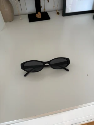 Svarta smala solglasögon - Snygga svarta solglasögon med smal, oval form och helsvart båge. Perfekta för en trendig och stilren look. Glasen är mörka och bågarna är i plast, vilket gör dem lätta och bekväma att bära.