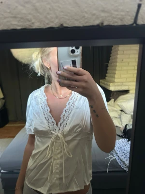 Vit blus med spets och knytband - Söt vit blus med djup V-ringning dekorerad med spetskant. Knytband i midjan och små knappar framtill. Kortärmad modell i ett lätt, glansigt material.