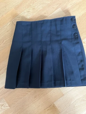 Svart plisserad omlottkjol kortkjol med knappar - Snygg svart omlottkjol kortkjol med plisserade veck och tre dekorativa knappar upptill i sidan. Kjolen har en klassisk och stilren look som passar till många olika outfits.