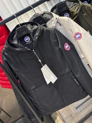 Svart vindjacka från Canada Goose - Snygg svart vindjacka från Canada Goose med huva och dragkedja framtill. Jackan har en klassisk logotyp på ärmen och praktiska fickor. Perfekt för blåsiga dagar och enkel att matcha med olika outfits.