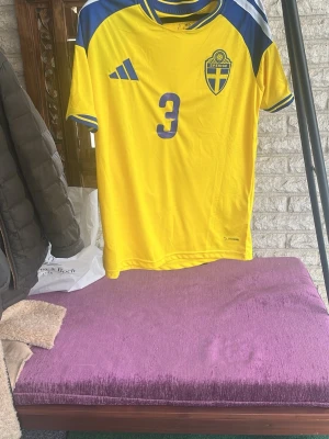 Sverige fotbollströja Lindelöf #3 - Gul och blå  Sverige fotbollströja med Lindelöf och nummer 3 på ryggen. Tröjan har korta ärmar, rund hals och blå detaljer på axlar och ärmslut. Perfekt för fotbollsfans som vill stötta landslaget. Jag säljer den för att den var för stor för mig