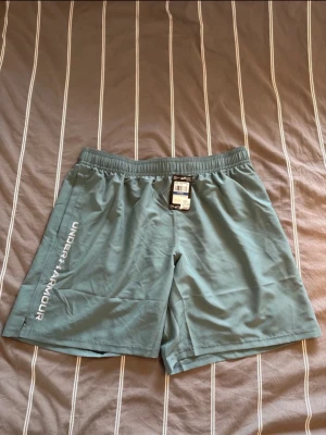 Gröna träningsshorts från Under Armour - Snygga gröna  träningsshorts från Under Armour, oanvända med prislapp. Nypris: 379kr  med vit logga längs benet. Shortsen har elastisk midja och är tillverkade i ett lätt och snabbtorkande syntetmaterial. Perfekta för gymmet eller löprundan.