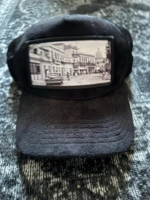 Svart keps med vintage stadsmotiv - Svart keps med böjd skärm och mesh på baksidan. Framtill finns en stor patch med ett vintage svartvitt stadsmotiv. Kepsen har klassisk trucker-modell och justerbar passform. Perfekt för dig som gillar unik streetstyle. Väldigt bra skick, hör av dig om du har flet frågor eller vid intresse av köp!