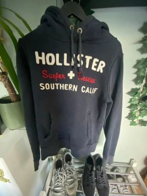 Hollister hoodie - Mörkblå hoodie från Hollister med broderat vitt och rött tryck på bröstet där det står 'Hollister Surfer Rescue Southern Calif'. Klassisk känguruficka och justerbar huva med snören. Perfekt för en avslappnad och cool stil.