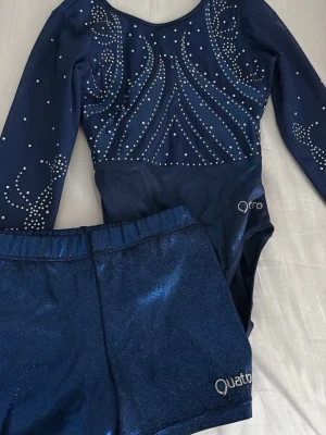 Blå gymnastikdräkt och shorts Quatro - Säljer en glittrig mörkblå tävlingsdräkt med långa ärmar och matchande shorts från Quatro. Dräkten har vita strasstenar i snygga mönster på bröstet och ärmarna. Perfekt för träning eller tävling inom gymnastik. Har används till 2 tävlingar så nästan helt ny. Köptes för ungefär 1500kr 