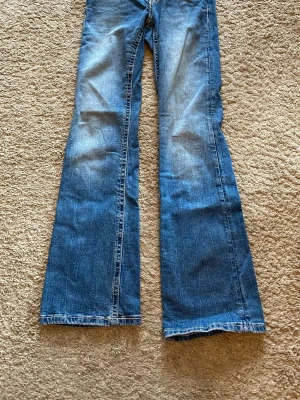 Blå bootcut jeans med slitningar - Säljer ett par blå bootcut jeans med snygga slitningar framtill och klassiska bakfickor med detaljer. Jeansen har en utställd passform nertill och är tillverkade i denim. Perfekta för dig som gillar retrostil och vill ha ett par jeans med lite extra attityd.