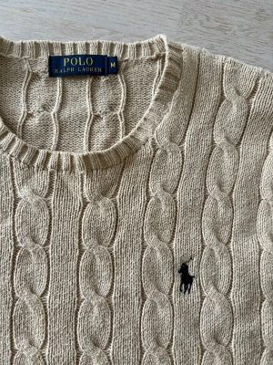 Beige kabelstickad tröja Polo Ralph Lauren - Kabelstickad beige tröja från Polo Ralph Lauren med rund halsringning och broderad logga på bröstet. Tröjan har långa ärmar och ribbade muddar vid ärmslut och nederkant. Perfekt för en stilren och klassisk look.