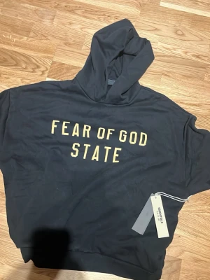 Svart hoodie Fear of God Essentials - Svart hoodie från Fear of God Essentials med texten 'FEAR OF GOD STATE' i gult framtill. Oversized passform och stor huva. Perfekt för en streetwear-look. Materialet känns mjukt och skönt mot huden.