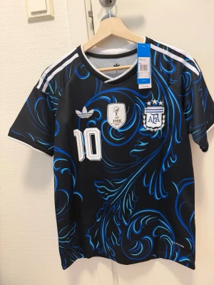 Argentina Messi tröja svart/blå Adidas - Snygg svart fotbollströja från Adidas med blått mönster, officiella Argentina-emblemet och nummer 10 Messi på ryggen. Tröjan har vita detaljer och klassiska Adidas-ränder på axlarna. Perfekt för dig som älskar fotboll och vill sticka ut på planen.