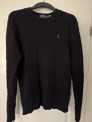 Svart Polo Ralph Lauren tröja - Kabelstickad svart tröja från Polo Ralph Lauren med rund halsringning och den klassiska röda logotypen broderad på bröstet. Tröjan har långa ärmar och ribbade muddar vid ärmslut och nederkant. Perfekt för dig som gillar stilren och tidlös design.