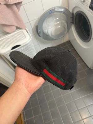 Svart keps från Gucci med logomönster - Snygg svart keps från Gucci med diskret GG-logomönster över hela kepsen. På sidan finns det klassiska gröna och röda bandet. Justerbar baktill för perfekt passform. Perfekt accessoar för en trendig streetstyle.