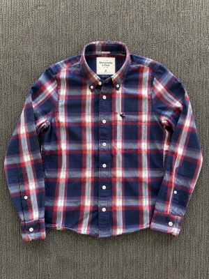 Rutig skjorta från Abercrombie & Fitch - Snygg rutig skjorta i blått, rött och vitt från Abercrombie & Fitch. Skjortan har klassisk krage, knappar framtill och långa ärmar. Storlek M.
