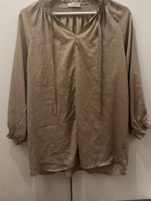 Beige blus med smockad halsringning - Snygg beige blus från Free/Quent med smockad halsringning och knytband. Blusen har långärmade ärmar med resår vid ärmslut och är tillverkad i ett lätt, glansigt material som ger en avslappnad men trendig look.