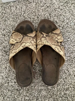 Slip-in sandaler med ormskinnsmönster - Coola slip-in sandaler med breda korslagda remmar i beige och brunt ormskinnsmönster från ATP Atelier. Sandalerna har öppen tå och låg klack, perfekt för en trendig och avslappnad look. Materialet är äkta läder med prägling av  ormskinn. Viss slitning i sulan - se foto. Nypris 2500 kr.