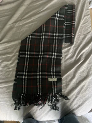 Rutig halsduk från Burberry i lammull - Snygg halsduk från Burberry i klassiskt rutigt mönster med svart, vitt och röda detaljer. Tillverkad i 100% lammull och har fransar i ändarna. Märkt med Burberry London och tillverkad i England. Perfekt accessoar för att lyfta din outfit.