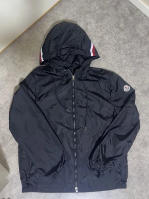 Svart Moncler vindjacka med huva - Snygg svart vindjacka från Moncler med huva och dragkedja framtill. Jackan har Moncler-logga på ärmen och vita samt röda detaljer på huvan. Justerbar dragsko i huvan och elastiska muddar vid ärmslut. Perfekt för en stilren och sportig look. Går att skanna!