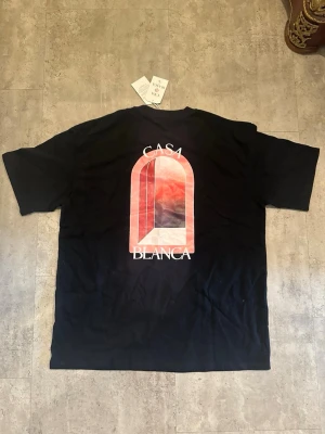 Svart Casa Blanca t-shirt med tryck - Cool svart t-shirt från Casa Blanca med ett stort grafiskt tryck i rött och vitt på ryggen och ett mindre tryck framtill. T-shirten har klassisk passform och rund hals. Perfekt för dig som gillar streetwear och vill sticka ut.