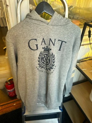 Grå hoodie från GANT med tryck - Snygg grå hoodie från GANT med stort marinblått tryck och emblem på bröstet. Klassisk huva och ribbade muddar. Perfekt för en avslappnad och sportig stil. Mjuk bomullsblandning som känns skön mot huden.