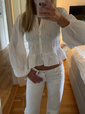 Vit blus med puffärmar och knappar - Trendig vit blus med långa puffärmar och knappar framtill. Blusen har smockdetaljer över bröstet och en lätt utsvängd nederkant. Tillverkad i ett tunt, luftigt tyg som ger en romantisk vibe. Perfekt att styla med jeans eller kjol.