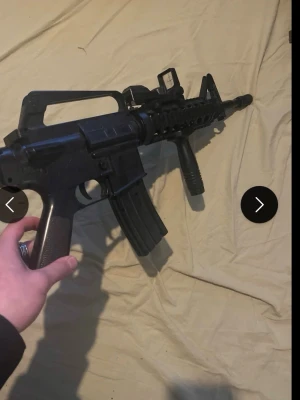 Svart airsoftgevär M15A1 - Säljer ett svart airsoftgevär i M4-stil med justerbar kolv, vertikalt grepp och sikte. Tillverkad i plast och metall, magasinet är löstagbart. Perfekt för airsoftspel och samling.
