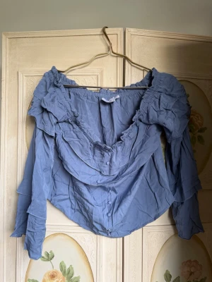 Blå volangblus med offshoulder - Snygg blå blus med offshoulder-design och volangdetaljer runt halsringningen och axlarna. Blusen har långa ärmar och en lös, avslappnad passform. Materialet känns lätt och luftigt.